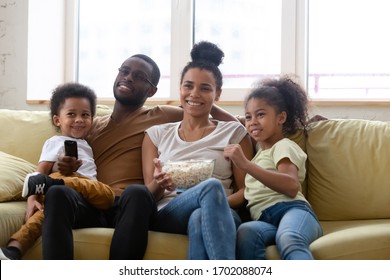 Happy African American Family And Children Watching Funny Tv Show Or Movie Eating Popcorn Snack. Happy Diverse Dad Holding Remote Controller, Mom Hugging Cute Siblings.