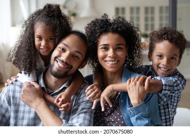 Happy African American family couple of parents piggybacking two kids at home, looking at camera with toothy smile. Sibling children hugging mom and dad. Parenthood concept. Head shot portrait