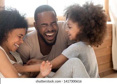 Happy african american family with cute little kid daughter tickling laughing at home together, cheerful black parents and small child girl having fun playing bonding enjoy funny lifestyle activity