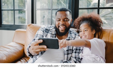 Happy African American Family Dad And Daughter Having Fun And Using Mobile Phone Video Call On Sofa At House. Self-isolation, Stay At Home, Social Distancing, Quarantine For Coronavirus Prevention.