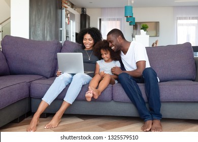 Happy African American Family With Daughter Using Laptop At Home, Shopping Online, Watching Video Or Movie, Smiling Mother And Father Spending Weekend Together With Child, Sitting On Sofa