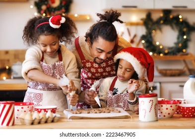 Happy African American Family Father And Two Kids In Festive Outfit Making Christmas Cookies Together In Kitchen At Home During Winter Holidays, Dad Decorating Xmas Gingerbreads With Small Children  