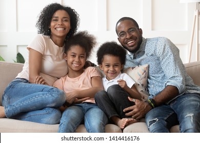 Happy african american family of four bonding looking at camera at home, smiling black parents and cute little kids sit on sofa, mixed race mom dad with small children embrace on couch, portrait