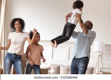 Happy African American Family Of Four Celebrating Moving In New Apartment. Black Smiling Father Lifting Small Son, Overjoyed Mommy Dancing With Laughing Daughter, Having Fun In Modern Living Room.