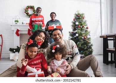 Happy African American Family With Gift Boxes Looking At Camera Near Blurred Grandparents And Christmas Tree