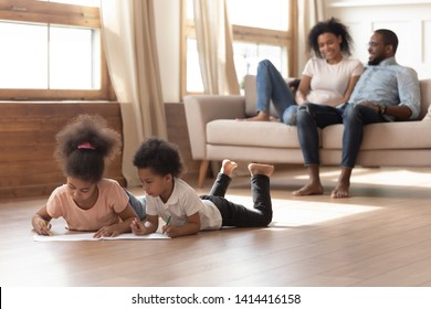 Happy African American Family With Kids Leisure Lifestyle Activities In Modern Living Room Concept, Young Black Parents Relax Talk Sit On Sofa Children Drawing Playing On Warm Wooden Floor At Home