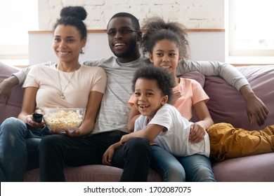 Happy African American Family With Kids Watching Funny Tv Show Or Movie Eating Popcorn Snack. Happy Diverse Dad Holding Remote Controller, Mom Hugging Cute Siblings.