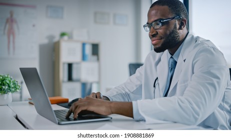 Happy African American Family Medical Doctor Is Working On A Laptop Computer In A Health Clinic. Physician In White Lab Coat Is Browsing Medical History Behind A Desk In Hospital Office.