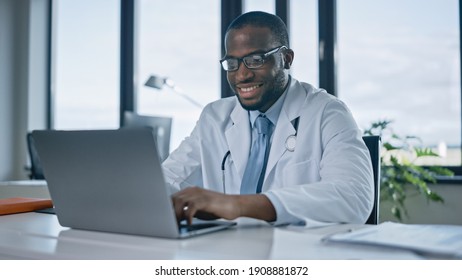 Happy African American Family Medical Doctor Is Working On A Laptop Computer In A Health Clinic. Physician In White Lab Coat Is Browsing Medical History Behind A Desk In Hospital Office.