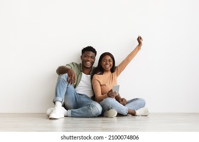 Happy African American Family Millennial Man And Woman In Casual Sitting On Floor At Empty Apartment, Using Mobile Phone Together, Lady Raising Hand Up And Smiling, Shopping Online, Copy Space