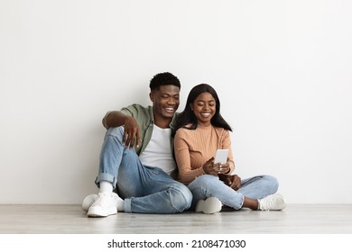 Happy African American Family Millennial Man And Woman In Casual Sitting On Floor At Empty Apartment, Using Mobile Phone Together, Shopping Online Or Websurfing On Internet, Copy Space