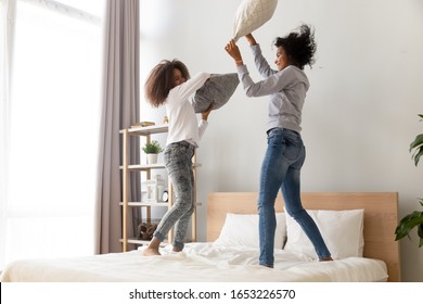 Happy african american family mom sister and teen daughter having fun pillow fight jumping on bed in the morning, excited black mother with teenager girl laugh play funny leisure activity in bedroom