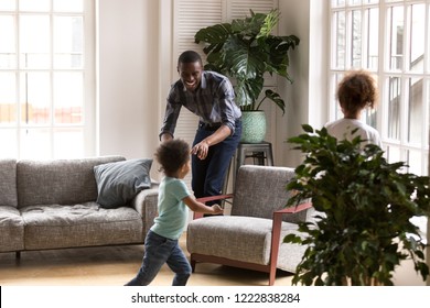 Happy African American Family Playing Together At Home, Laughing Father Playing Tag Touch Game With Little Preschooler Daughter And Toddler Son, Running In Living Room, Catching To Each Other