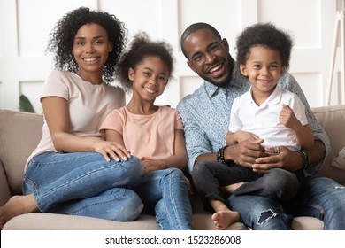 Happy African American Family Sitting On Couch Portrait. Smiling Dad Hold Preschool Son On Knees, Excited Black Mom Embracing Cute Daughter, Looking At Camera, Lazy Weekend Spending Together At Home.