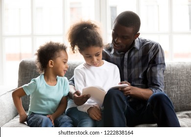 Happy African American Family Spending Time Together At Home, Focused Father Embracing Girl, Reading Fairytale Book, Textbook With Toddler Son And Teaching Preschooler Little Daughter To Read