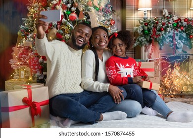 Happy African American Family Of Three Taking Selfie Near Christmas Tree At Home, Sitting On Floor Close To Fireplace In Decorated Living Room