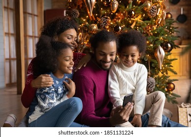 Happy African American Family With Two Kids Using Smartphone, Sitting Near Festive Tree At Home, Smiling Mother And Father With Son And Daughter Chatting Or Shopping Online On Christmas Holiday