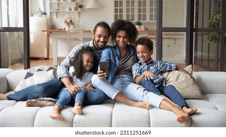 Happy African American family with two kids using smartphone, relaxing sitting on cozy couch, smiling mother and father with little son and daughter looking at phone screen, making video call - Powered by Shutterstock