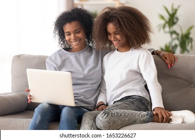 Happy African American Family Young Mother And Teen Daughter Using Laptop Computer Sitting On Couch, Smiling Mixed Race Parent Mom With Teenager Girl Having Fun Watching Movie Online At Home Together