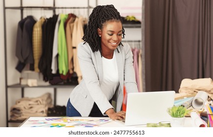 A happy African American fashion designer woman stands with her measuring tape in the studio, using computer, clothing spread out around her - Powered by Shutterstock