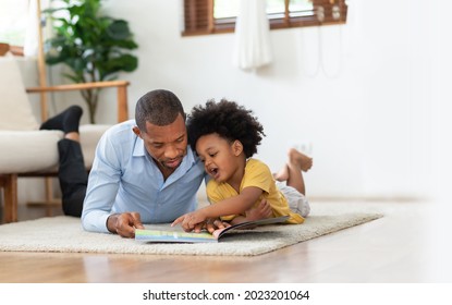 Happy African American Father And His Son Lying Reading Book On The Floor At Living Room.