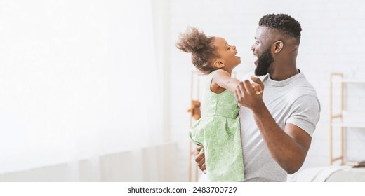 A happy African American father and his daughter are dancing together in a bright and spacious living room. They two are smiling and laughing as they enjoy their playful dance, copy space - Powered by Shutterstock