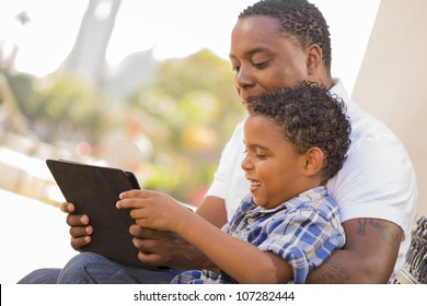 Happy African American Father And Mixed Race Son Having Fun Using Touch Pad Computer Tablet Outside.