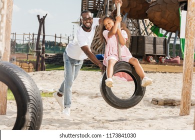 Happy African American Father Pushing Daughter On Tire Swing At Amusement Park