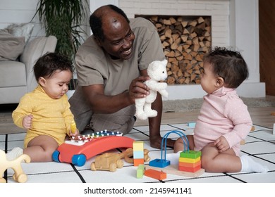 Happy African American Father Sitting On Floor In Living Room At Home Playing With His Cute Twin Babies Showing Them Soft Toy