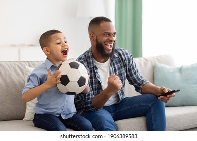 Happy African American Father And Son Watching Sports On TV Holding Soccer Ball And Remote Control Sitting On Sofa At Home. Family Watches Television. Sport TV Programming Concept