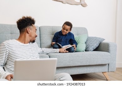 Happy African American Father And Son Reading Book And Using Laptop While Sitting At Home