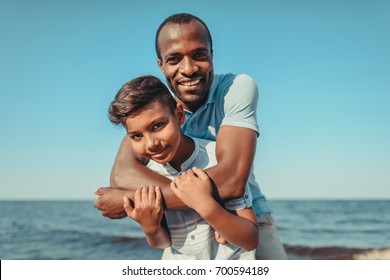 happy african american father and son smiling at camera while hugging on beach