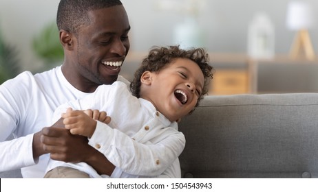 Happy African American father tickling laughing adorable son close up, family having fun at home, smiling dad holding hugging little boy, playing game, enjoying weekend, funny activity together