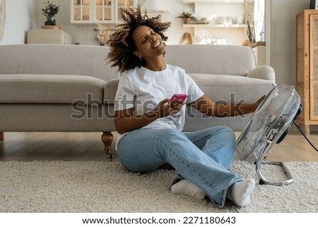 Happy african american female cools herself with electric ventilator rejoices in cool air in heat day. Smiling woman sit on floor with smartphone hold regulates airflow from home fan with flowing hair