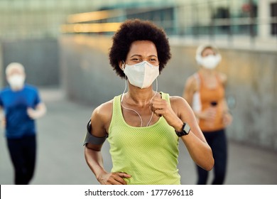 Happy African American female runner wearing protective face mask while jogging outdoors during coronavirus epidemic. There are people in the background. 