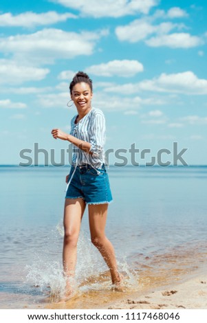 happy african american female tourist walking in sea water near sandy beach