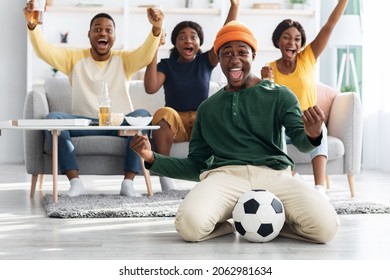 Happy african american football fans millennial men and women in casual watching match on TV at home, raising hands up and screaming, black guy with soccer ball over his emotional friends