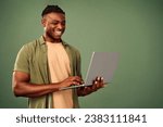 Happy african american freelancer in casual khaki shirt looking on laptop screen while typing on keyboard. Positive handsome man standing over green background and surfing internet.