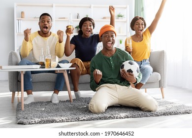 Happy African American Friends Watching Football Match Together At Home, Sitting On Couch In Living Room, Holding Football Ball, Screaming And Gesturing, Celebrating Success, Goal