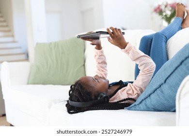 Happy african american girl lying on couch, listening to music and using tablet at home, copy space. Childhood, expression, technology, communication and domestic life, unaltered. - Powered by Shutterstock