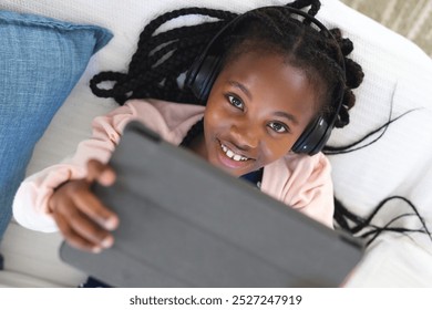 Happy african american girl lying on couch, listening to music and using tablet at home, copy space. Childhood, expression, technology, communication and domestic life, unaltered. - Powered by Shutterstock