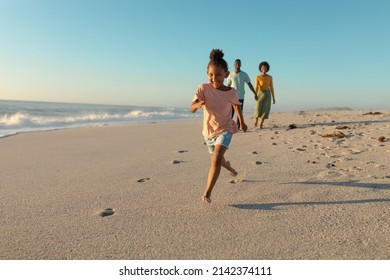 Happy African American Girl Running On Sand Against Parents Walking At Beach. Unaltered, Family, Lifestyle, Togetherness, Enjoyment And Holiday Concept.