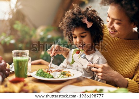 Happy African American girl sitting on mother's lap while she is feeding her at dining table.
