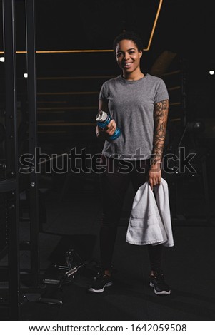 happy african american girl with tattoo holding sports bottle with water and standing in gym