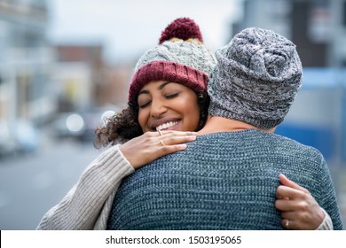 Happy African American Girlfriend Hugging Boyfriend On Street With Closed Eyes. Friends Meeting And Embracing Outdoor. Romantic Multiethnic Young Couple Embracing In Winter Clothes In The City.