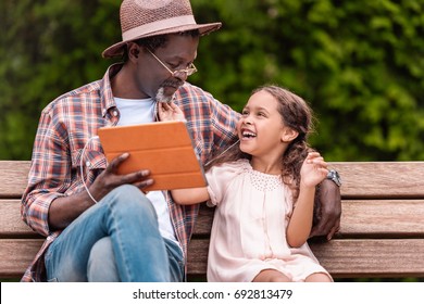 Happy African American Grandchild And Her Grandfather Listening Music On Digital Tablet While Sitting On Bench In Park  