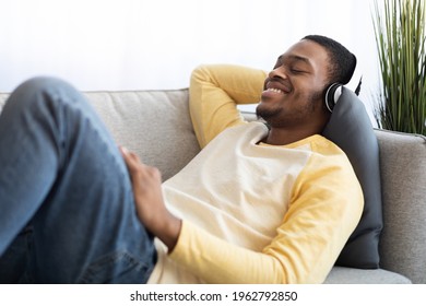 Happy African American Guy Enjoying Music, Using Headset, Side View. Smiling Young Black Man With Closed Eyes Listening To Music While Chilling Alone At Home, Using Brand New Headphones