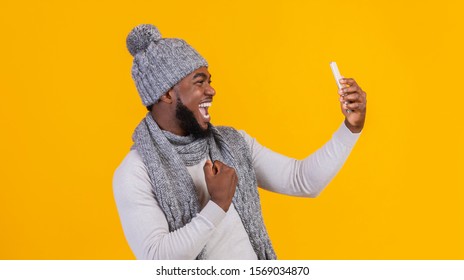 Happy African American Guy In Grey Winter Hat Taking Selfie On Phone Over Yellow Background, Side View