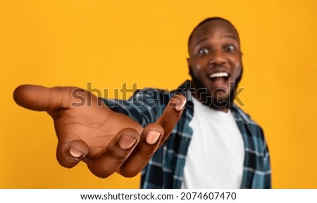 Happy african american guy outstretching hand, holding something invisible on his huge palm, giving it away or asking for something, orange studio background, collage, panorama with copy space