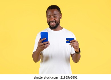 Happy African American Guy Shopping On Smartphone Holding Credit Card Standing Over Yellow Studio Background. Ecommerce And Mobile Banking Application Concept